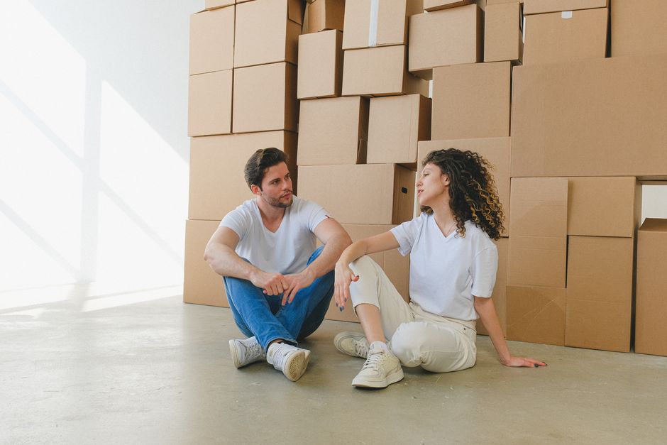 A man and a woman sitting on the floor inside a property during a home relocation process, with numerous cardboard moving boxes stacked against a white wall behind them. The man has dark hair and a be