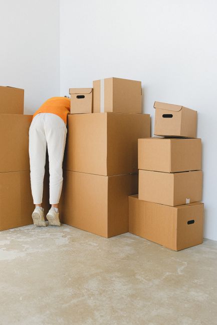 A man and a woman sitting on the floor inside a property during a home relocation process, with numerous cardboard moving boxes stacked against a white wall behind them. The man has dark hair and a be