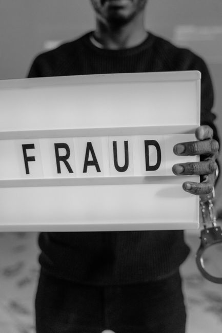 A person is holding a white lightbox sign with the word 'FRAUD' spelled out in black letters, gripping the sides with both hands. The individual's face is partially visible, with a blurred background