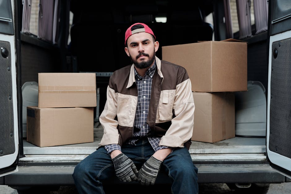 A young man with light brown hair and a beard, dressed in a blue zip-up hoodie over a white T-shirt and dark blue trousers, is sitting on the edge of an open delivery van during a home relocation proc