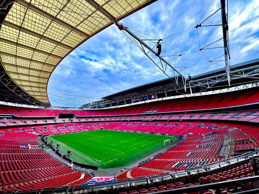 Interior view of Wembley Stadium featuring a lush green football pitch at the centre, surrounded by multiple tiers of red seating arranged in circular and curved formations. The stadium's roof structure, composed of metal and translucent panels, extends overhead, with visible supporting cables and trusses. The sky above shows a mix of blue sky and scattered clouds, casting natural light into the spacious interior. In the foreground, the loading area includes a flat pavement with a few personnel and equipment indicating ongoing relocation or delivery activities. Nearby, cardboard boxes, plastic wrapping, and moving blankets are visible, suggesting packing and furniture transport preparations related to house removals or home relocation services. The scene reflects a professional moving environment, with the space prepared for the transport of furniture and goods in connection with comprehensive moving logistics carried out by [COMPANY_NAME], supporting efficient packing, loading, and transport of property belongings.