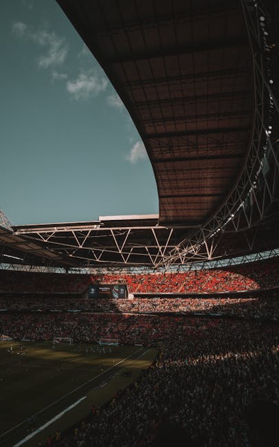 Inside a large stadium with a partially open roof structure, showing the expansive seating area filled with spectators and a field below. The roof's metal framework and roofing material are clearly visible, with sunlight streaming in through the open sections, illuminating the interior. The perspective looks upward towards the roof, capturing the scale and architecture of the venue. This setting represents a large-scale event at a well-known sports stadium, suitable for house removals and relocation services involving transport and logistics for major sporting or entertainment venues, as part of the broader moving and packing process handled by companies like Man and Van Brent.