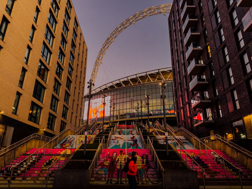 Interior view of Wembley Stadium featuring a lush green football pitch at the centre, surrounded by multiple tiers of red seating arranged in circular and curved formations. The stadium's roof structure, composed of metal and translucent panels, extends overhead, with visible supporting cables and trusses. The sky above shows a mix of blue sky and scattered clouds, casting natural light into the spacious interior. In the foreground, the loading area includes a flat pavement with a few personnel and equipment indicating ongoing relocation or delivery activities. Nearby, cardboard boxes, plastic wrapping, and moving blankets are visible, suggesting packing and furniture transport preparations related to house removals or home relocation services. The scene reflects a professional moving environment, with the space prepared for the transport of furniture and goods in connection with comprehensive moving logistics carried out by [COMPANY_NAME], supporting efficient packing, loading, and transport of property belongings.
